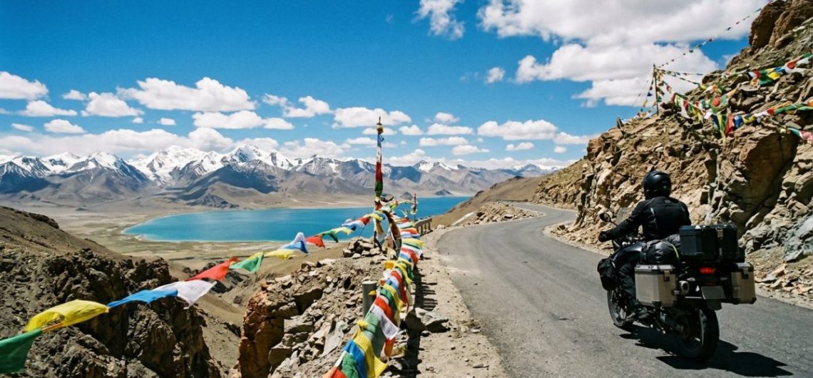 Touring motorcycle on a high Tibetan plateau road with prayer flags, snow peaks and a turquoise lake